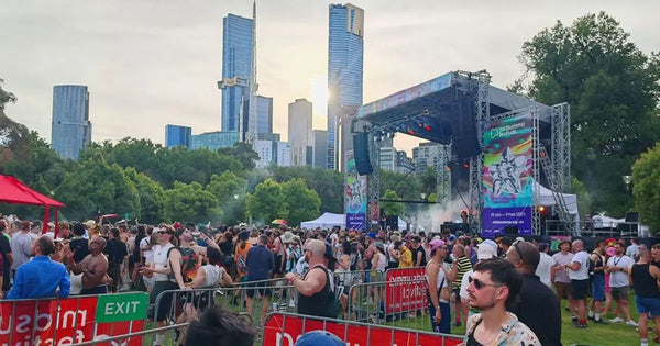 Attendees enjoying the Midsumma Festival in a park setting, with the Melbourne skyline and vibrant stage banners enhancing the festive atmosphere.