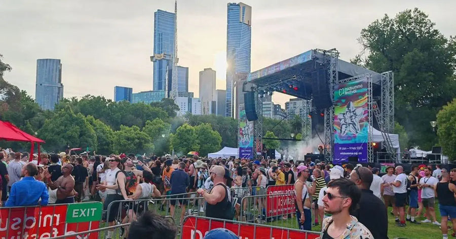 Attendees enjoying the Midsumma Festival in a park setting, with the Melbourne skyline and vibrant stage banners enhancing the festive atmosphere.