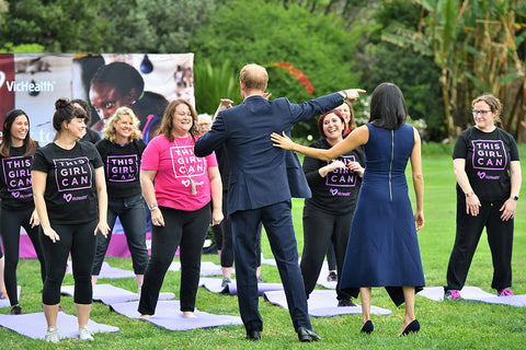 A group of people outdoors on a grassy lawn, including Prince William and Princess Mary, standing on yoga mats and wearing black T-shirts printed with the slogan 'THIS GIRL CAN' in white and pink. Several women in the background are wearing bright pink T-shirts with the same slogan.