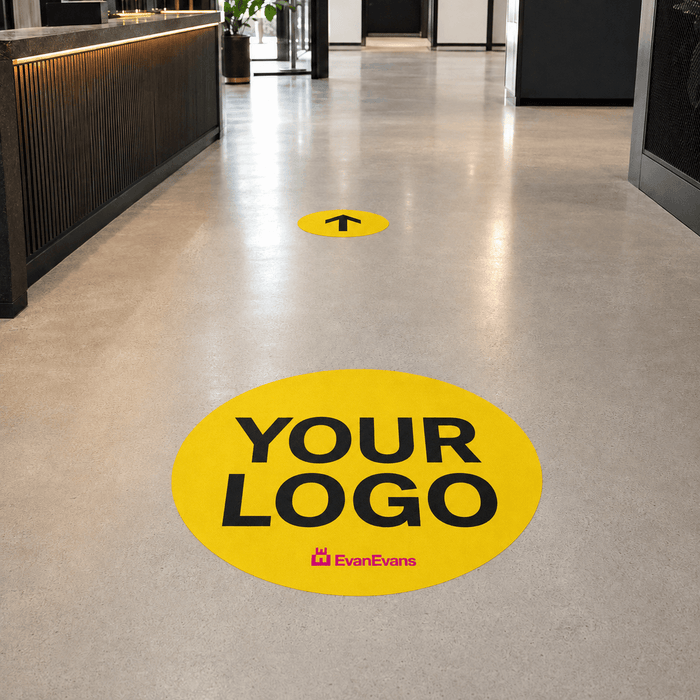 A bright yellow circular floor decal with black text reading 'YOUR LOGO' and a small red EvanEvans logo, placed on a polished gray office floor in a modern lobby. A smaller matching yellow arrow sticker is visible further down the hall.