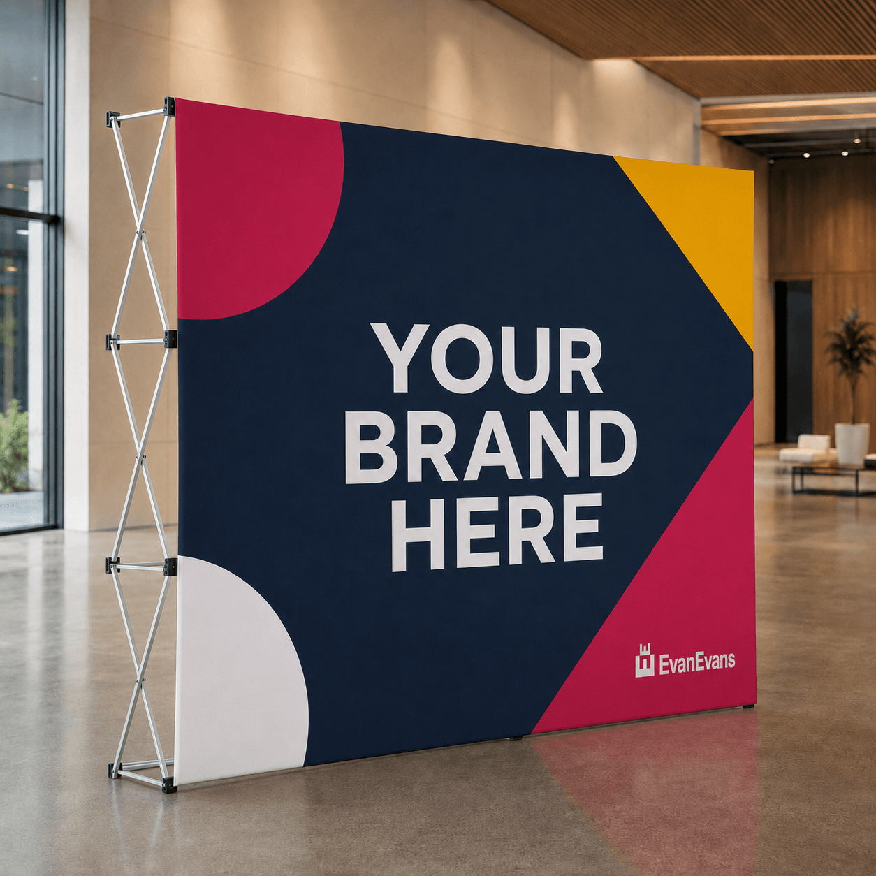 A freestanding rectangular banner stand with a large printed backdrop in a modern indoor lobby. The backdrop has abstract geometric shapes in navy, magenta, yellow, and white, with the text 'YOUR BRAND HERE' and a small 'EvanEvans' logo near the lower right.