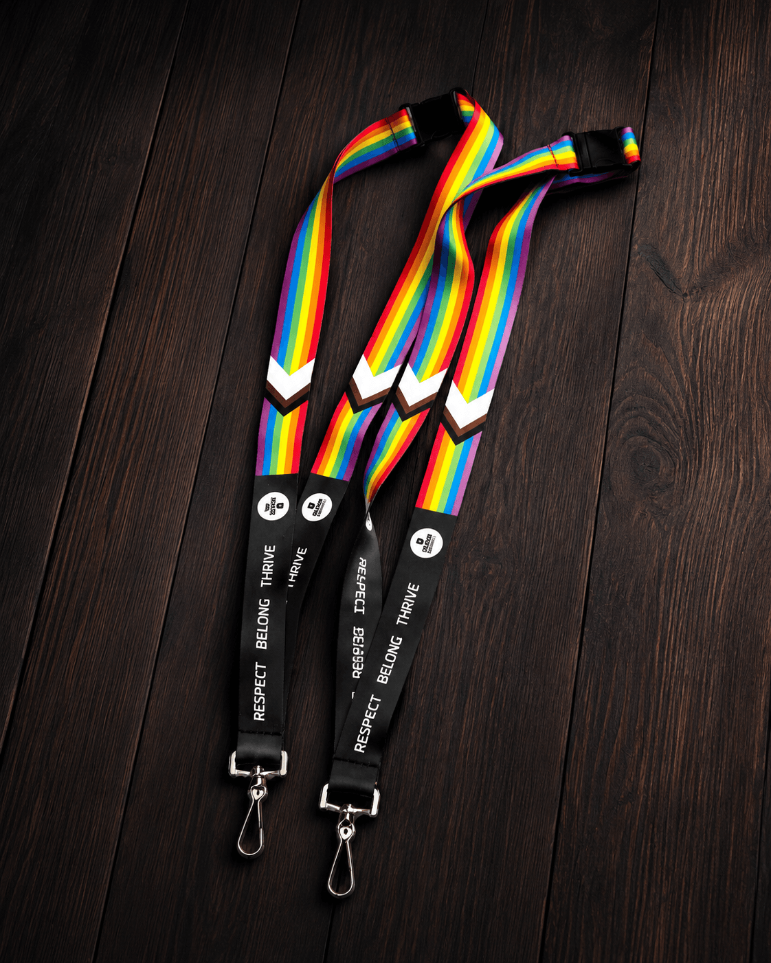 Two rainbow-striped lanyards with black webbing and silver metal clips, laid flat on a dark wood background. The black sections include white text reading 'RESPECT BELONG THRIVE' and circular white logos.