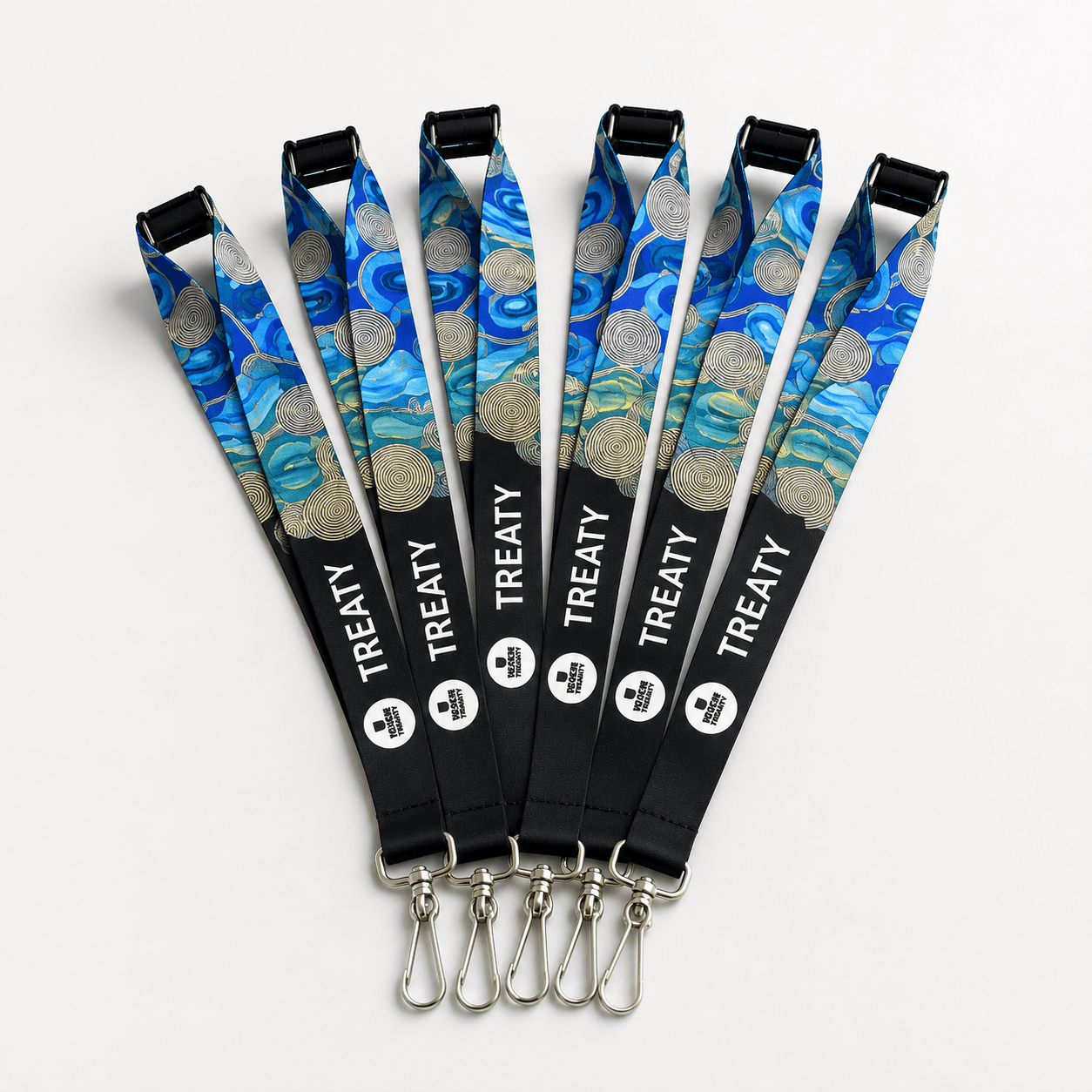 Six lanyards arranged in a fan shape on a light gray background. Each lanyard has a blue and teal abstract print with gold coin motifs near the top, a black lower section, the word 'TREATY' in white, and silver metal clip hardware.