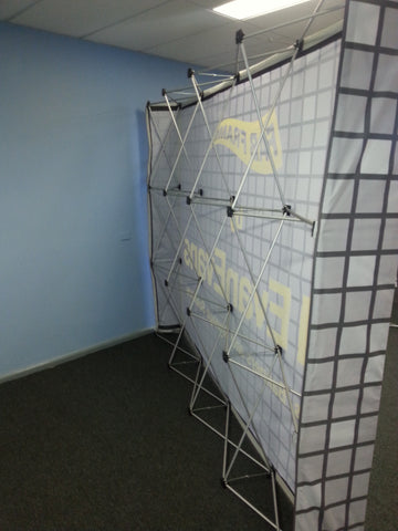 A freestanding modular display wall or backdrop with a white grid-patterned fabric panel, black framing, and yellow printed text, set in an indoor room with blue walls and dark carpet.