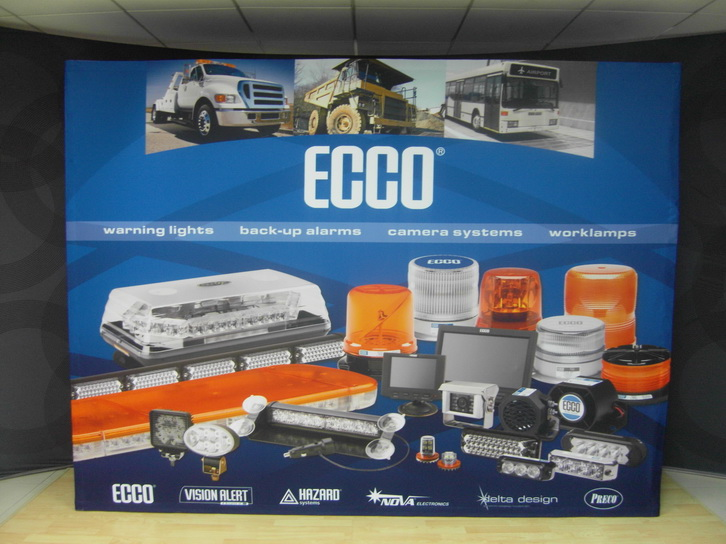 A large blue ECCO signage and lighting products display board featuring various commercial vehicle warning lights, amber and white dome lights, camera systems, and worklamps, photographed in an indoor setting.
