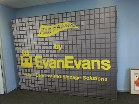 A large printed banner or backdrop with a gray grid pattern and bold yellow text reading 'EvanEvans' and 'Flags, Banners and Signage Solutions,' photographed leaning against a blue wall in an indoor room.