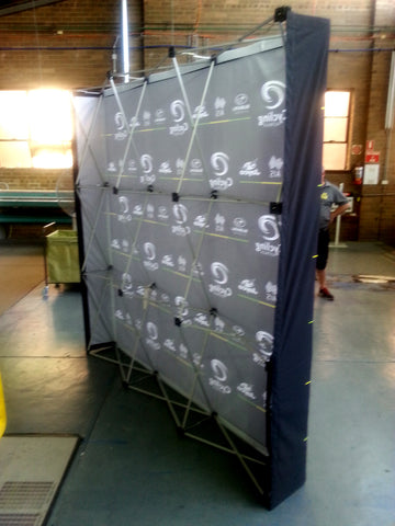 A freestanding folding banner or backdrop with a dark gray printed front panel covered in repeated white logos, supported by a metal frame, photographed indoors in a warehouse-like space.