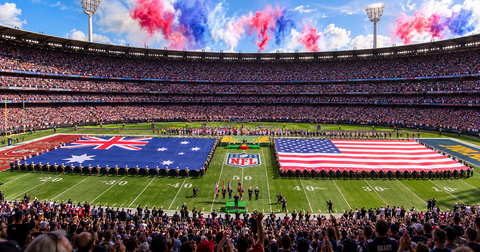 A wide stadium scene showing a football field filled with large patriotic flag formations, a crowd in the stands, and red, white, and blue smoke in the sky. The NFL logo is visible at center field.