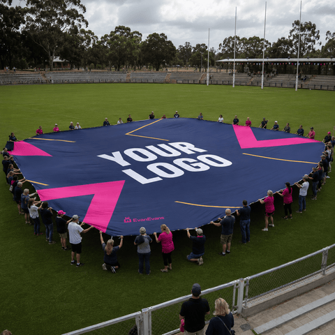 A large custom printed blue banner or mat is held in a circle by many people on a green sports field. The banner features a dark blue background with large pink geometric shapes and white text reading 'YOUR LOGO'.