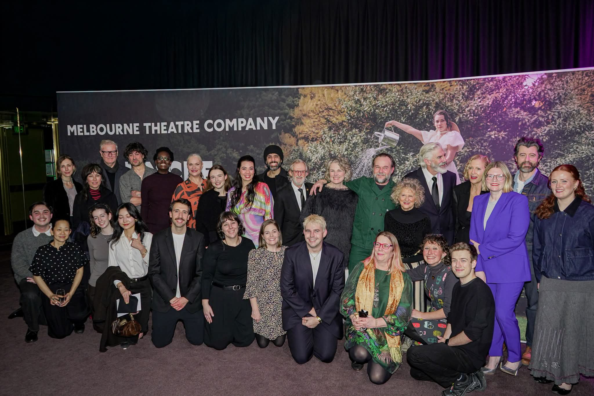 A group of people posing in front of a large 'Melbourne Theatre Company' backdrop banner at what appears to be an event or premiere.