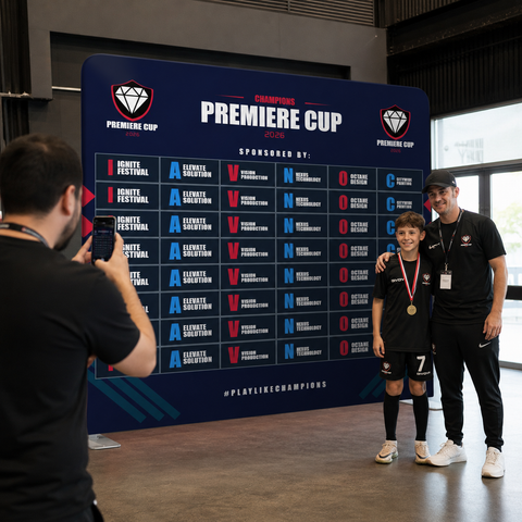 Two people posing in front of a 'Premiere Cup' backdrop with a young person holding a medal.