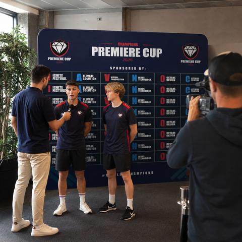 Two men in sports attire standing in front of a 'Premiere Cup' backdrop with a cameraman filming them.